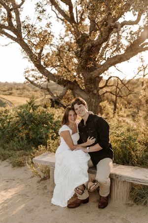 Couple sitting on a wooden bench under a large tree at golden hour, woman in white dress, man in black shirt.