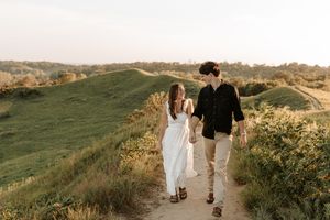 A couple walks hand-in-hand along a scenic hillside path at golden hour, woman in white dress, man in black shirt.