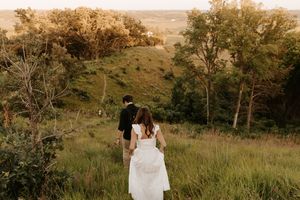 Bride and groom walking hand in hand through lush green meadow at golden hour during outdoor wedding.