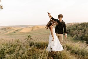 Couple dancing outdoors in a golden field, man spinning woman in white dress with rolling hills in background.