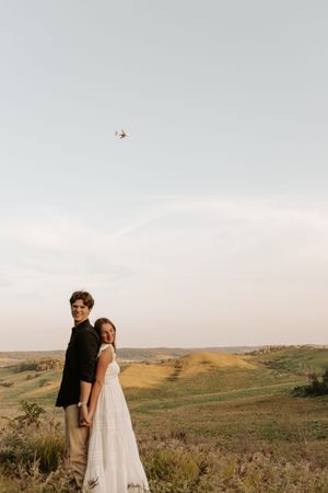 Couple standing back-to-back on a scenic hilltop in golden fields, woman in white dress, drone visible in sky.
