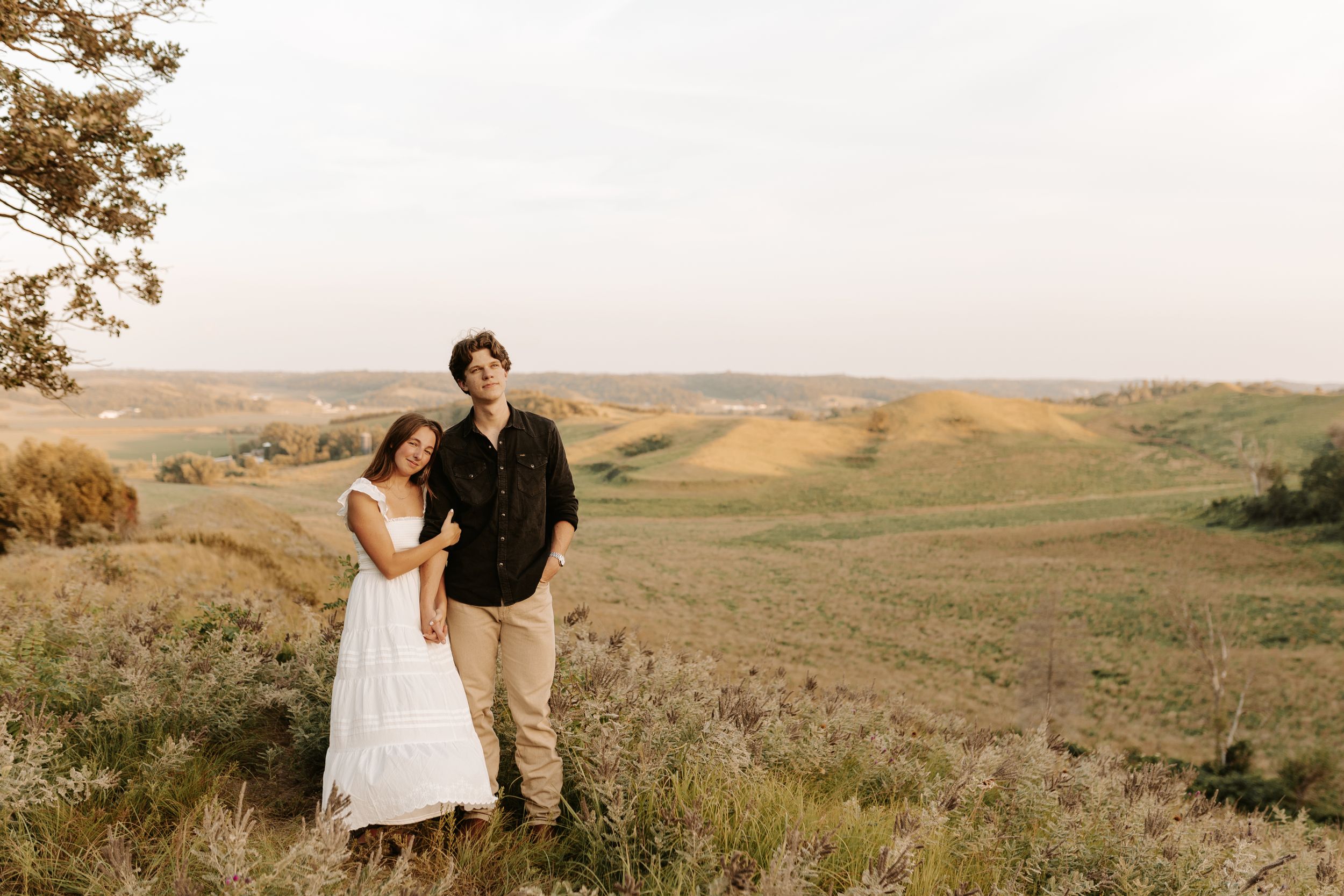 Couple posing together on a scenic hillside with rolling green fields and golden grass in the background at sunset.