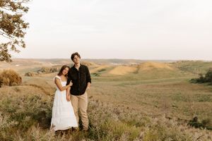 Couple posing together on a scenic hillside with rolling green fields and golden grass in the background at sunset.