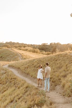 Couple holding hands on a winding dirt path through golden fields at sunset, woman in white dress holding bouquet.