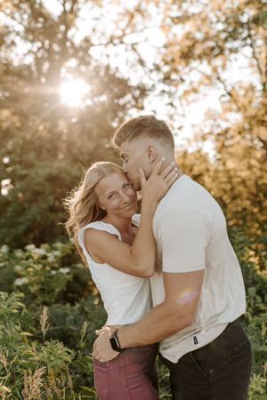 Couple embracing outdoors at golden hour, woman smiling at camera while man kisses her cheek among lush greenery.