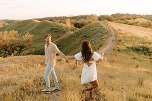Couple holding hands dancing in golden field with rolling hills and dirt path in background at sunset.