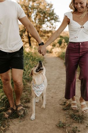 Couple holding hands with a white Lab wearing a floral bandana on a scenic dirt trail.