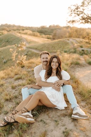 A couple sits together on a sandy hillside at golden hour, woman in white dress, man in light blue jeans, smiling warmly.