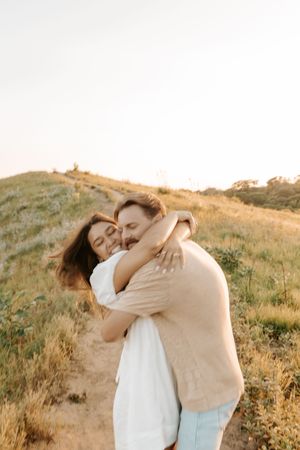 Happy couple embracing on a sunny hillside path, smiling warmly in golden hour light with green meadows behind them.
