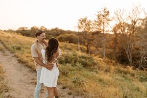 Couple embracing and smiling at golden hour on a scenic dirt path surrounded by golden grass and trees.