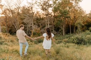 Couple holding hands walking through golden meadow with trees at sunset.
