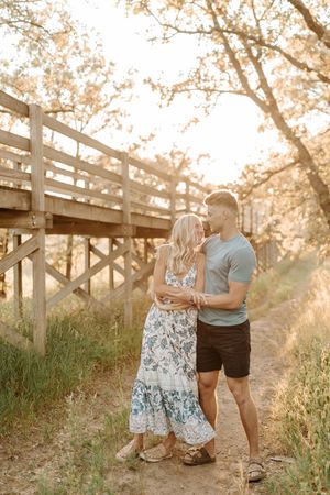 Couple sharing a romantic moment on a sunlit dirt path beside a wooden bridge surrounded by golden trees.
