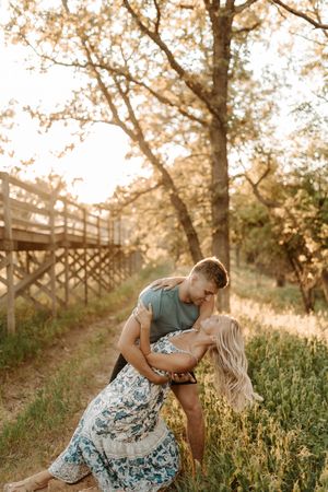 Couple sharing a romantic dip kiss on a sunlit path surrounded by golden trees and wildflowers.