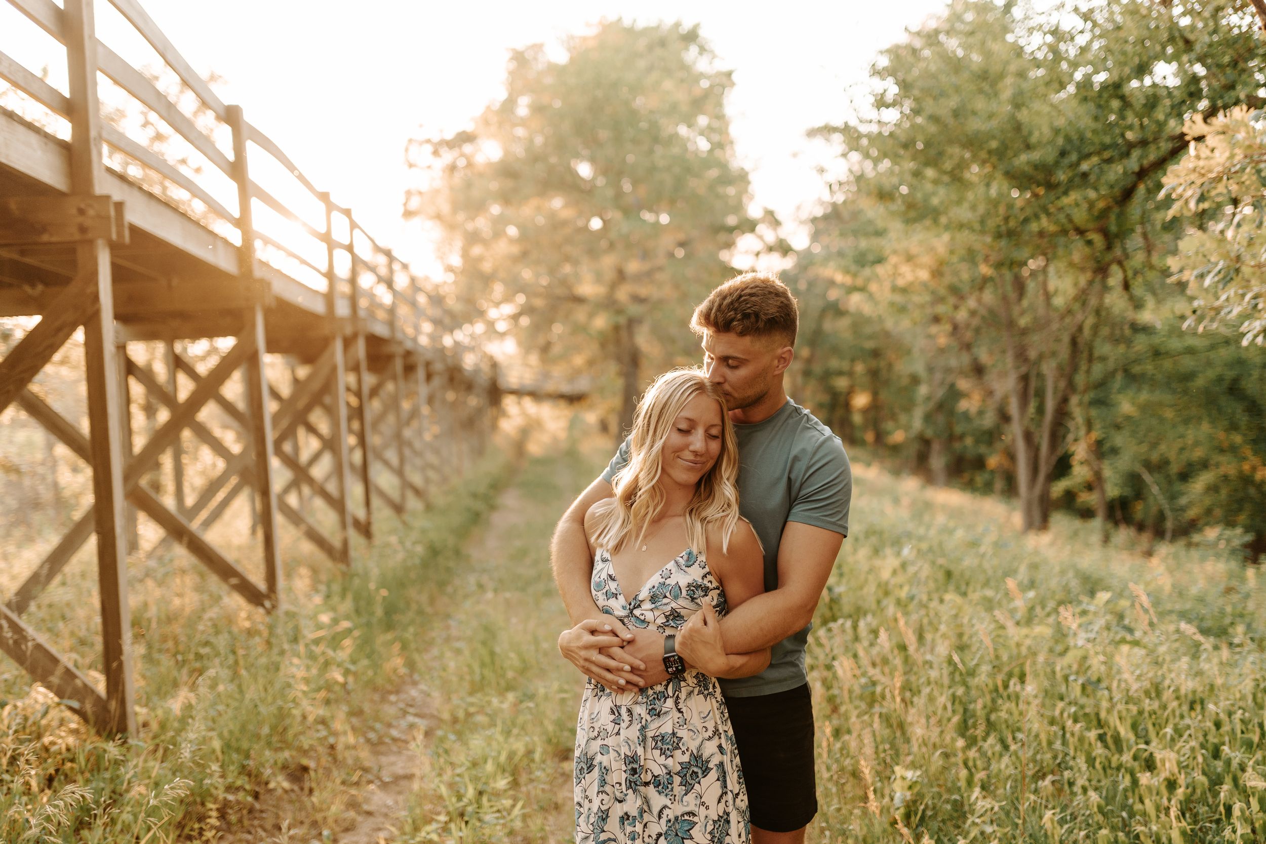 Couple embracing outdoors near wooden bridge surrounded by lush greenery in warm golden sunset light.