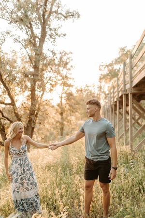 A couple holds hands in a sunlit meadow near a wooden bridge, the woman in a floral maxi dress and man in a teal tee.