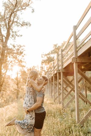 Couple embracing and kissing at golden hour near wooden stairs surrounded by lush greenery and warm sunlight.