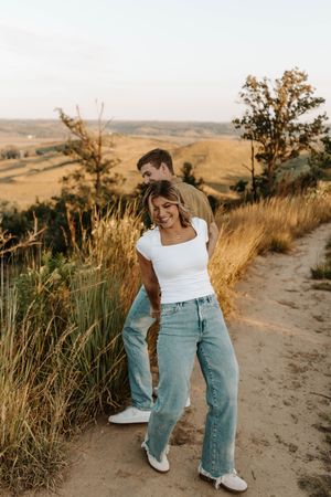 Couple embracing on a scenic overlook trail, woman in white tee and jeans smiling, golden grasslands in background.
