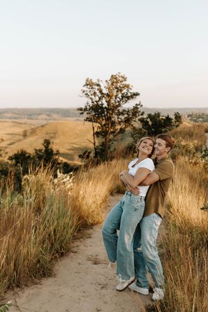 Happy couple embracing on a scenic overlook trail surrounded by golden grasslands and rolling hills at sunset.