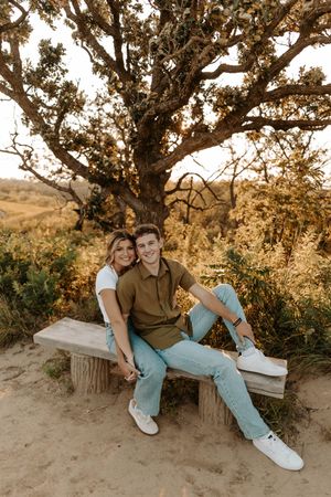 A couple sits together on a rustic bench under a golden tree, smiling warmly in a scenic outdoor setting.
