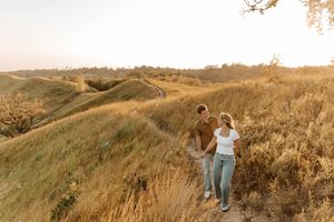 Couple holding hands while hiking golden grass hills at sunset in a romantic outdoor engagement photo.