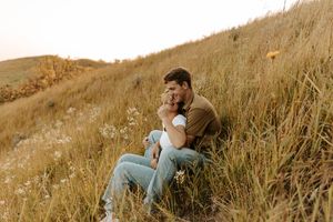 Couple sitting together in golden grass field on a hillside, sharing a tender moment during a romantic outdoor photo session.
