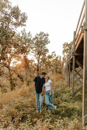Couple sharing a romantic kiss outdoors at golden hour near a wooden cabin surrounded by autumn foliage.