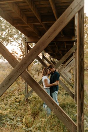 Couple embracing romantically under a wooden bridge structure surrounded by lush greenery in a fall outdoor setting.