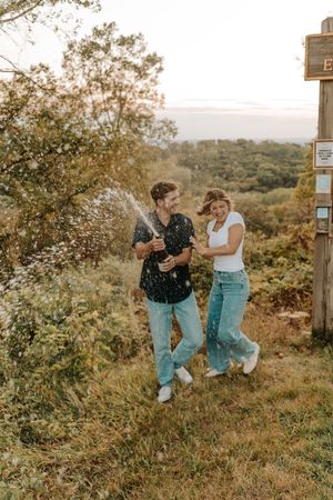 Couple celebrating outdoors by popping champagne bottle, wearing jeans and casual tops, surrounded by lush greenery.