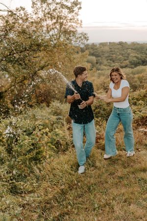 Couple celebrating outdoors by popping champagne bottle, laughing joyfully in a scenic green landscape.
