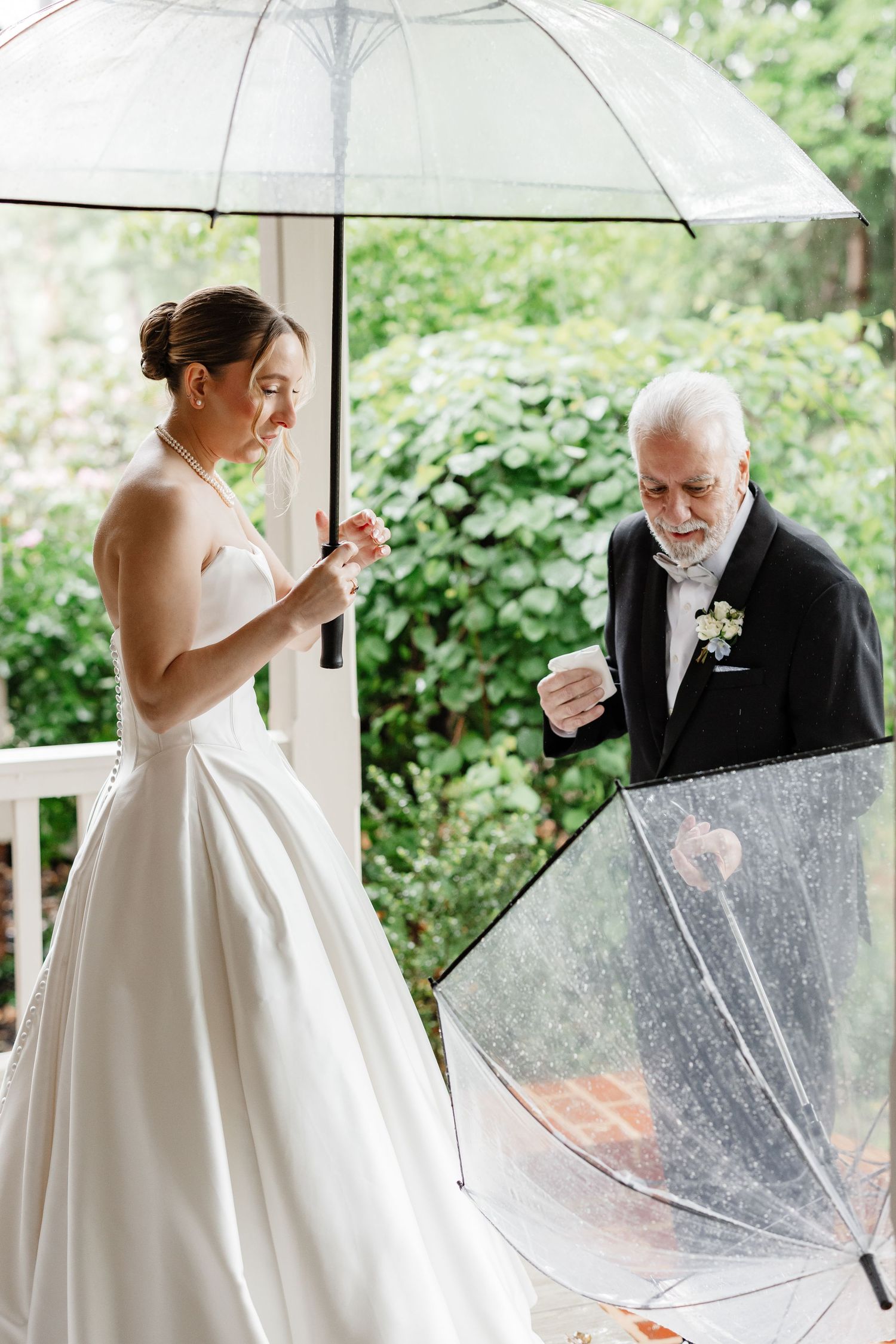 Bride in strapless ball gown opens clear umbrella with older gentleman in tuxedo on rainy wedding day.