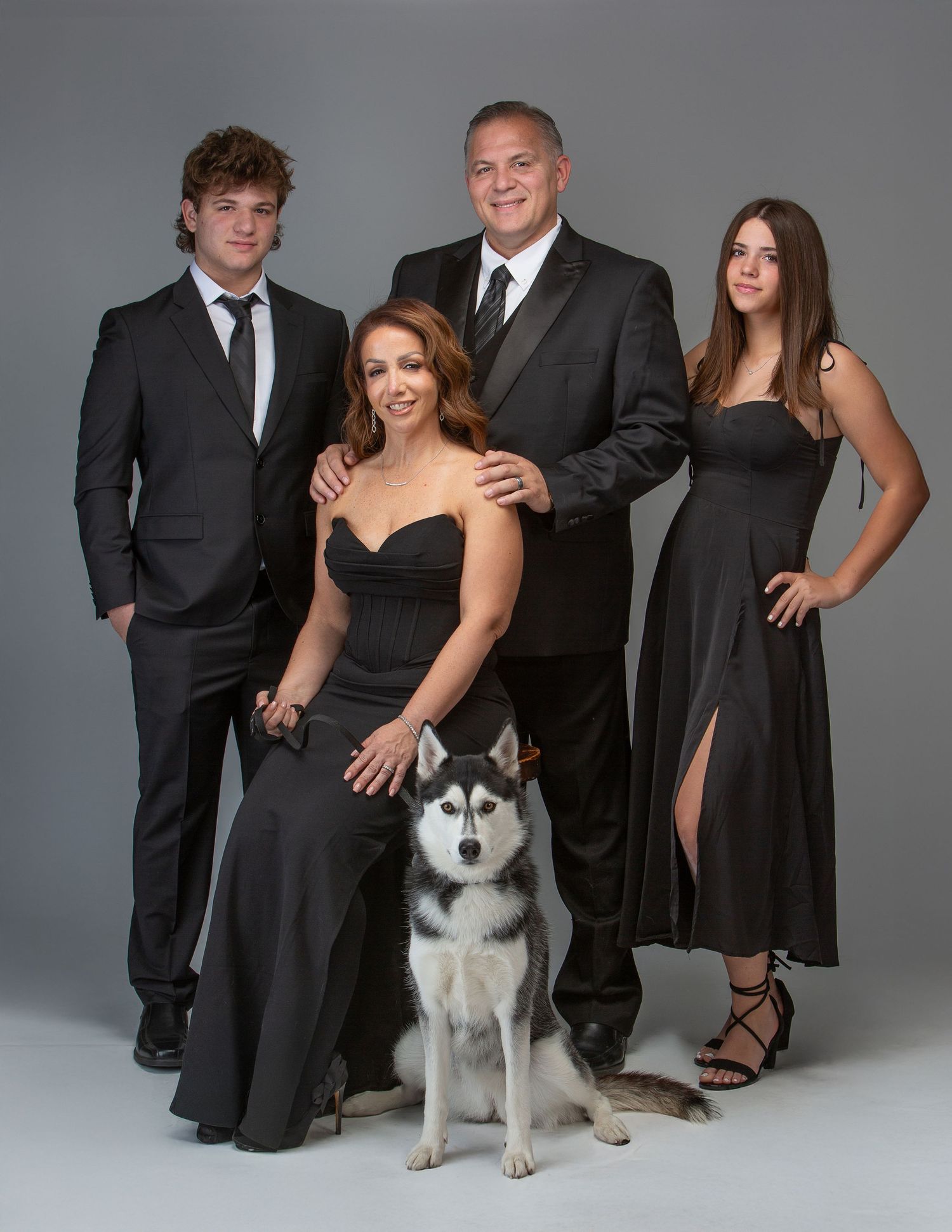 Family of four in elegant black attire posing with a Husky dog in a professional studio portrait.