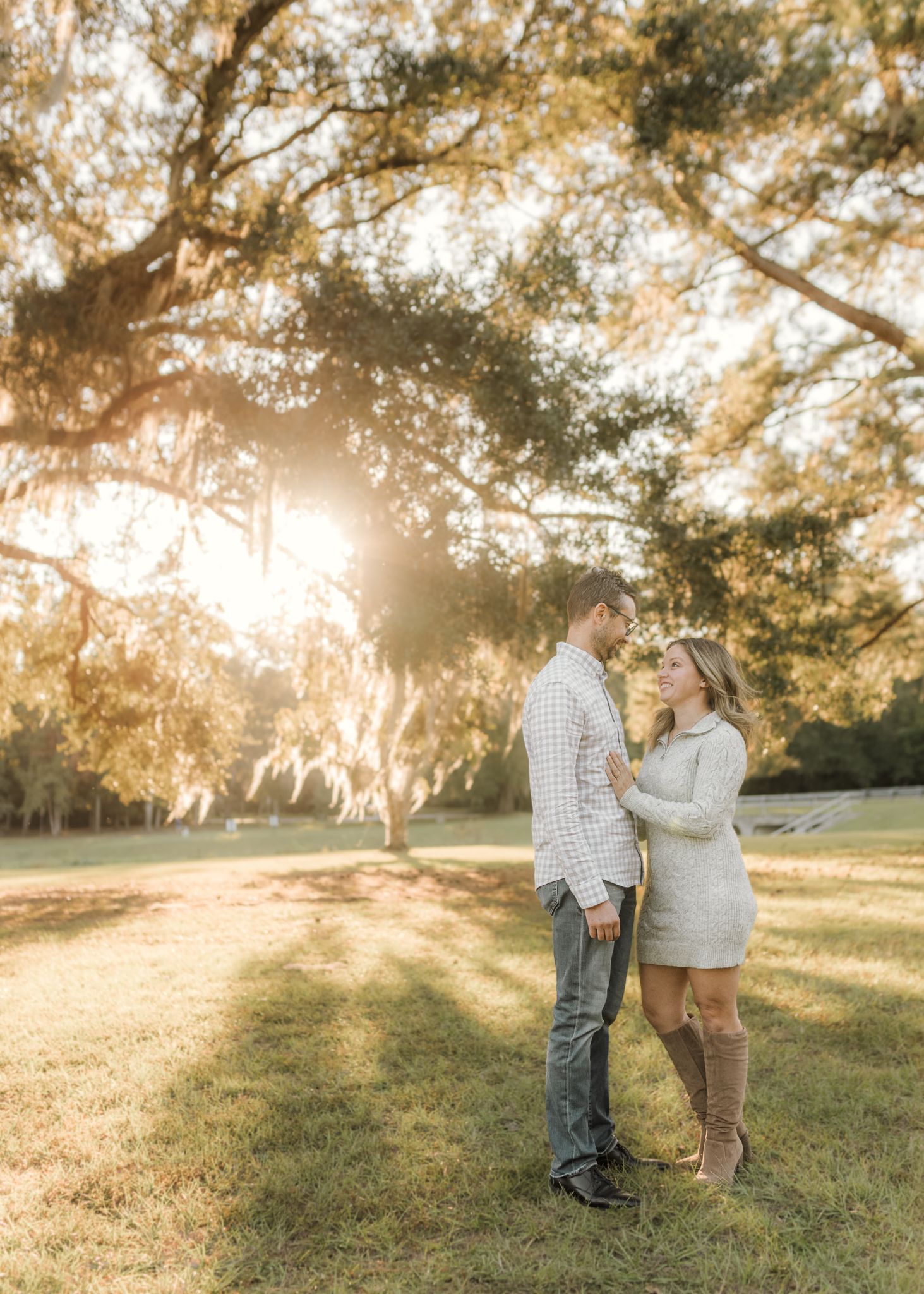 Couple standing together under golden oak trees at sunset, woman in cream sweater dress and brown boots, man in plaid shirt.