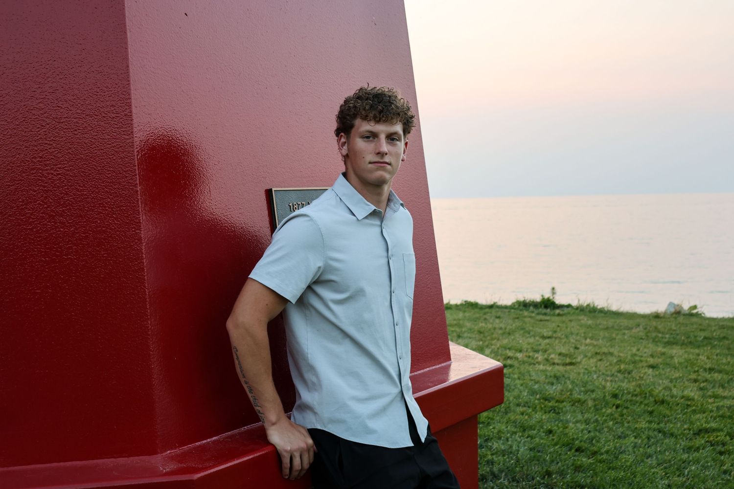 Young man with curly hair in a light shirt leaning against a red structure near the ocean at sunset.