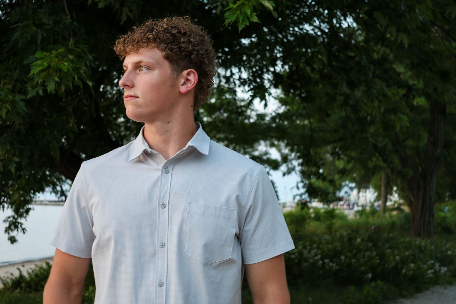 Young man with curly hair in a light button-up shirt looking away outdoors surrounded by green trees.