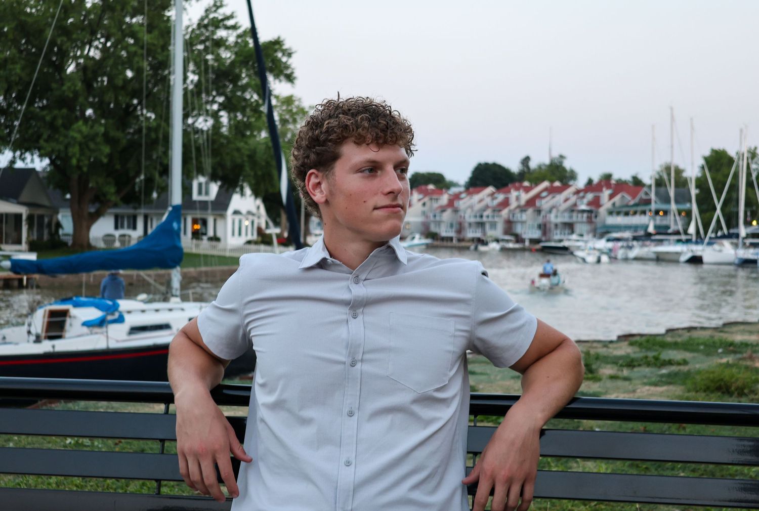Young man with curly hair in a light shirt posing by a bench at a scenic marina at dusk.