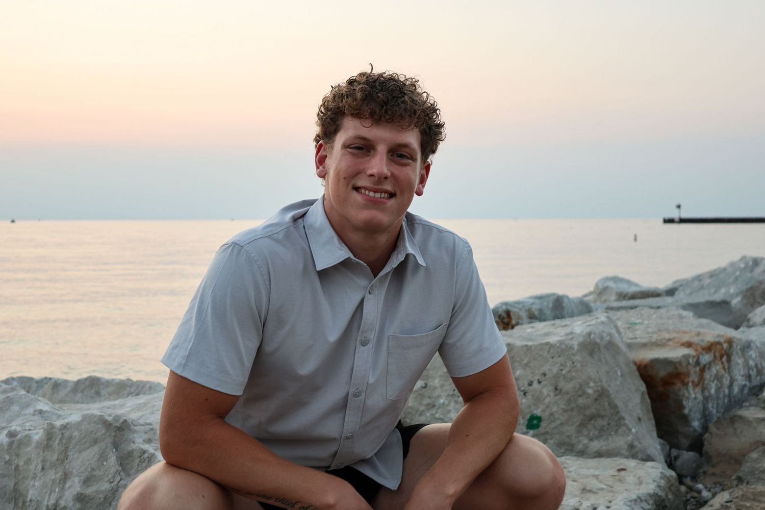 Smiling young man with curly hair sitting on rocks by the ocean during a colorful sunset.