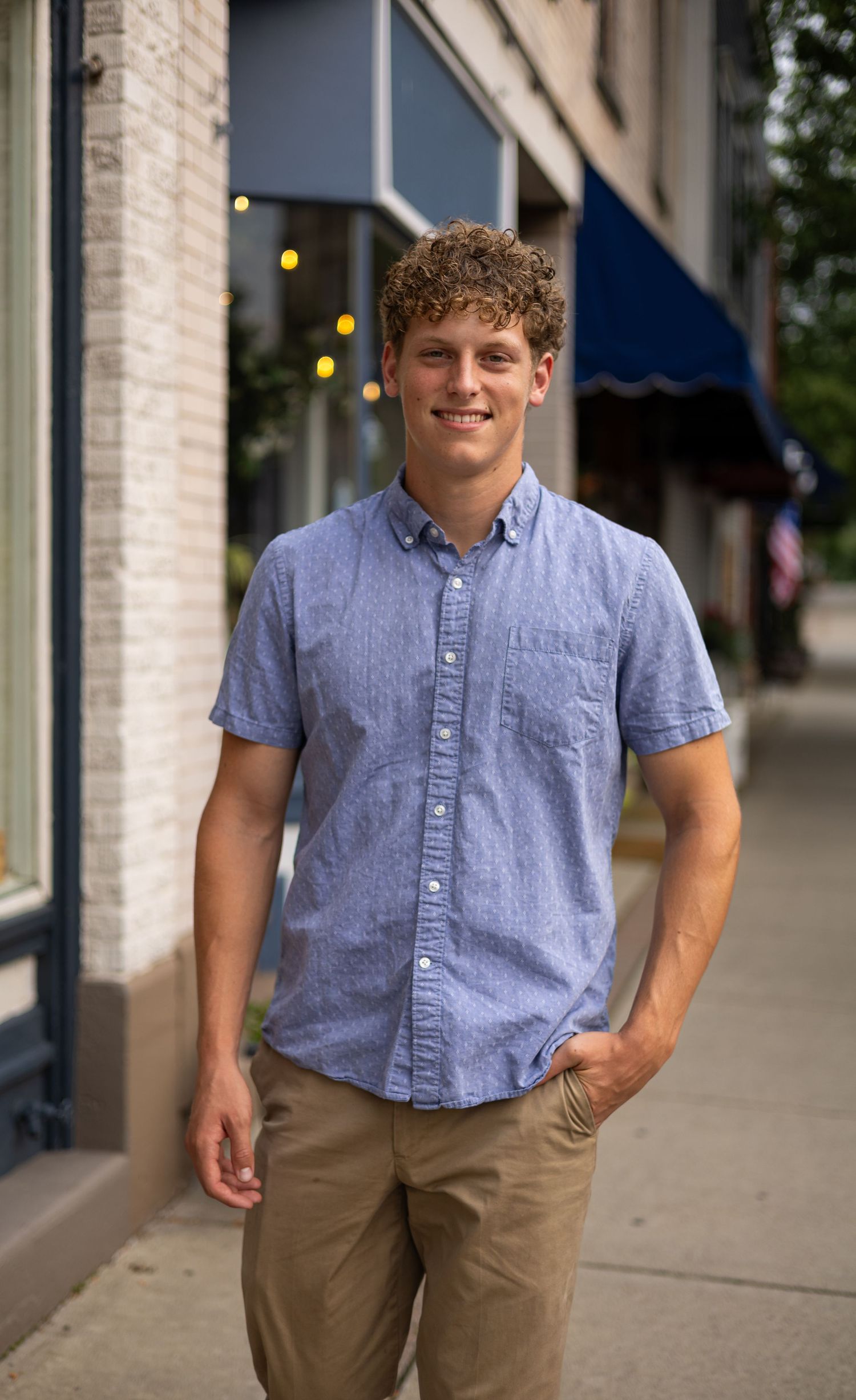 Smiling young man with curly hair in a blue button-up shirt standing on a sunny urban sidewalk.