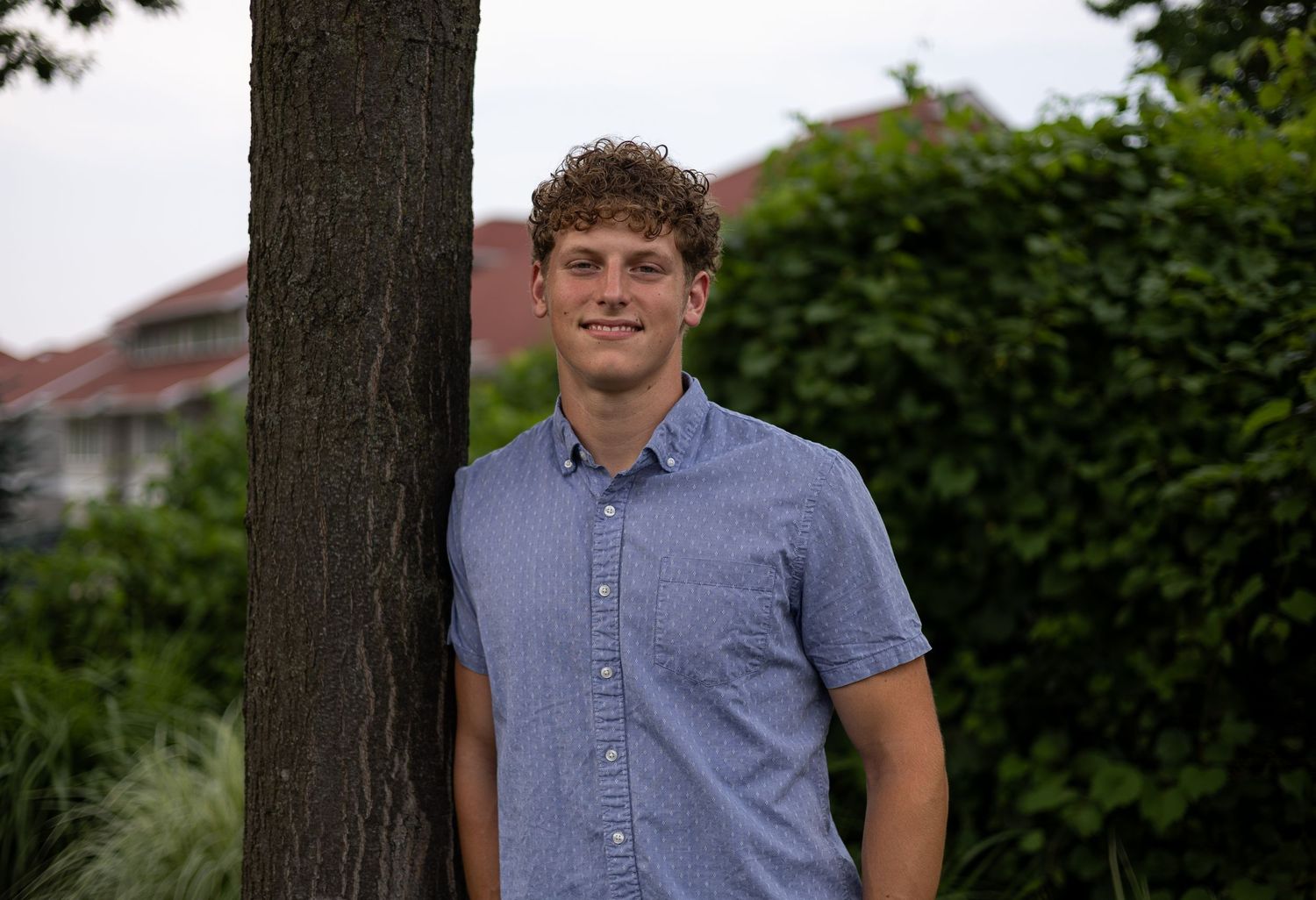 Smiling young man with curly hair in a blue shirt leaning against a tree in a lush green garden.