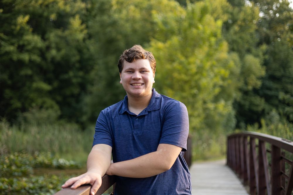 Smiling young man in blue polo shirt leaning on wooden bridge railing surrounded by lush green trees.