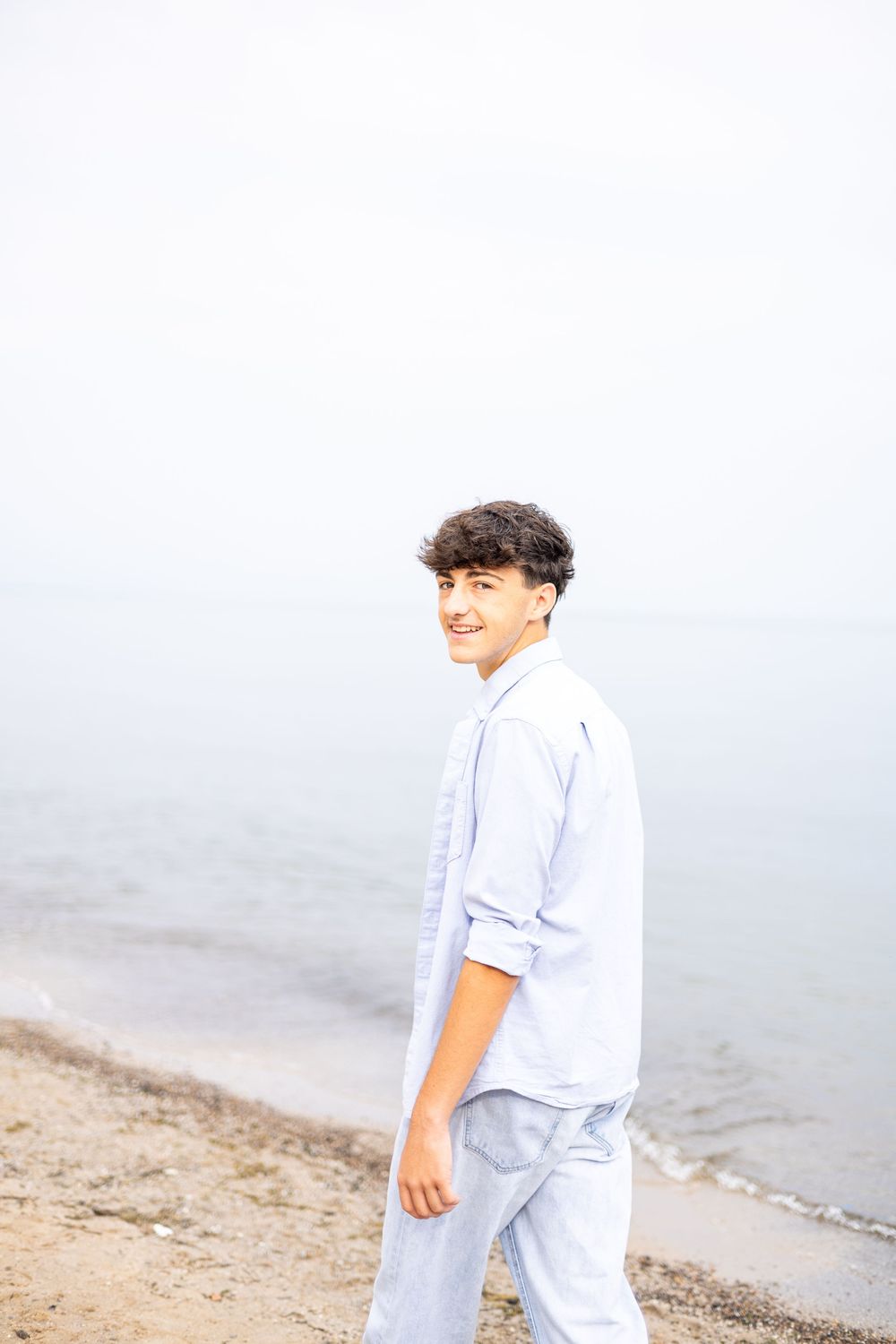 A smiling teenage boy in a light blue shirt and pants walks along a sandy beach with calm water in the background.