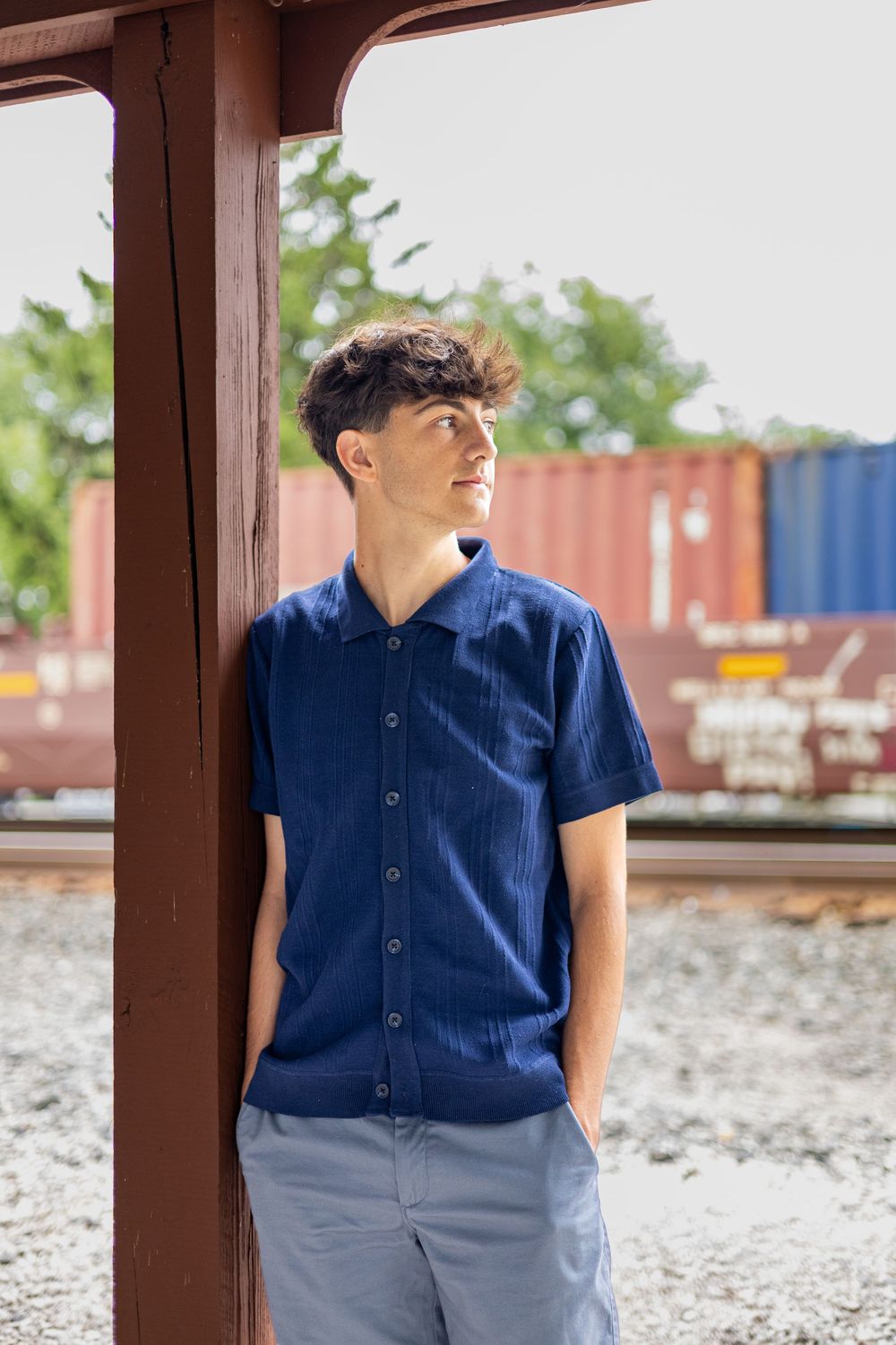 Young man in blue button-up shirt leaning against a rusty pole near railroad tracks with cargo containers in background.