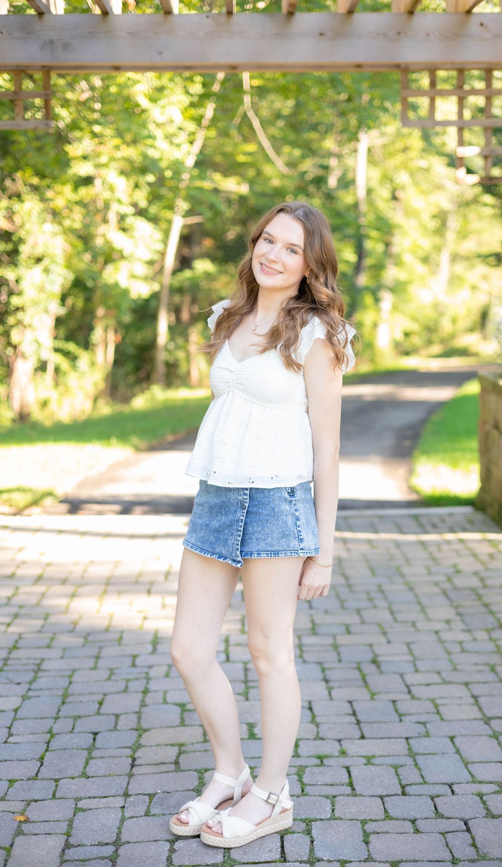 Young woman smiling in white top and denim skirt with wedge sandals, standing on stone path in lush green park.
