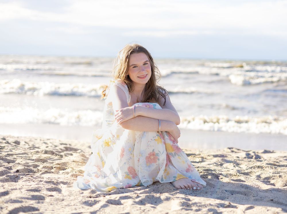 Young woman in floral dress sitting on sandy beach, smiling with ocean waves in background.