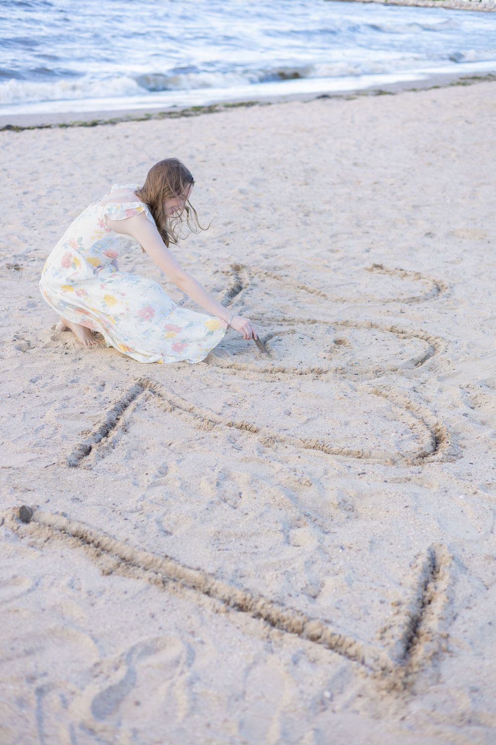 Young girl in floral dress writing numbers in beach sand near ocean waves on a sunny day.