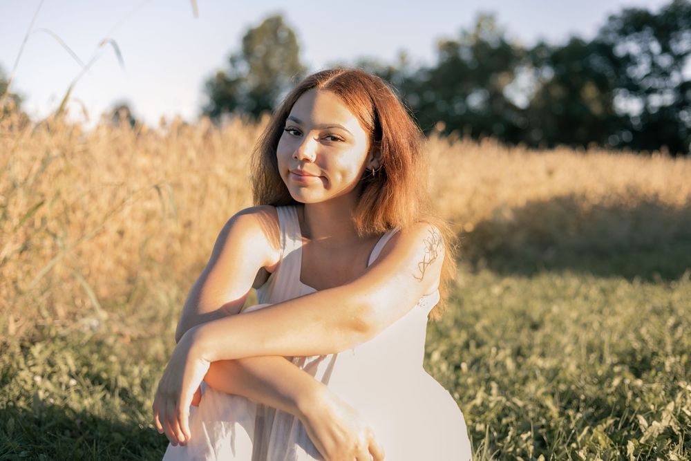 Young woman in white dress sitting in golden grass field, smiling warmly in soft sunlight.