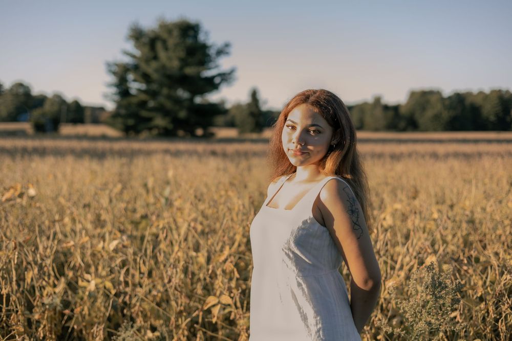 Young woman in white dress smiling in golden field at sunset with trees in background.