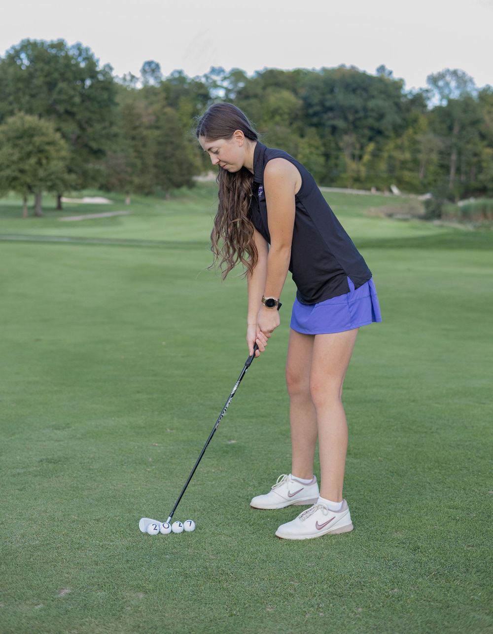Young female golfer in blue skirt and black top practicing her putting stance on a green golf course.
