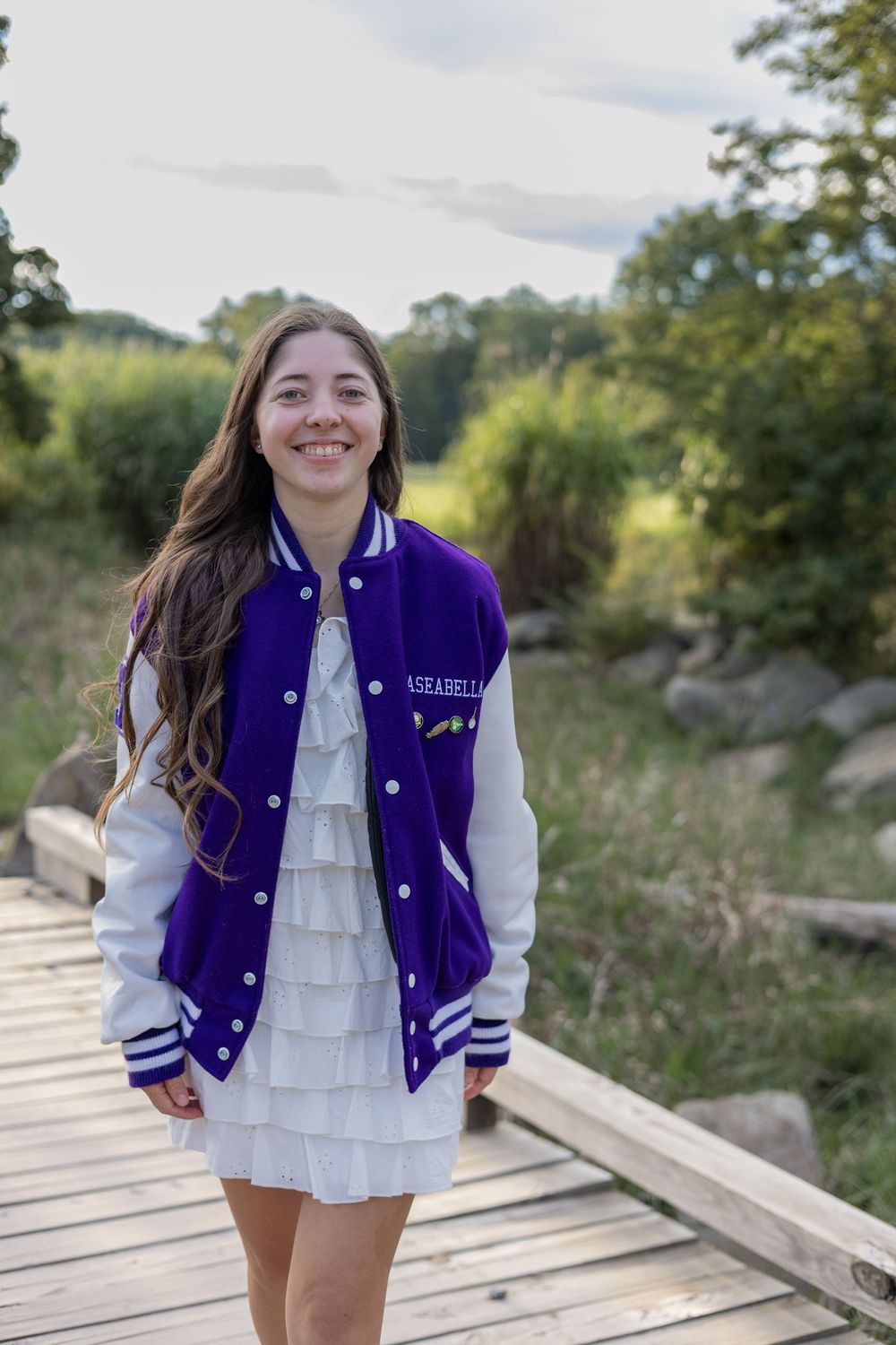 Smiling young woman in purple varsity jacket and white dress standing on wooden bridge in nature.