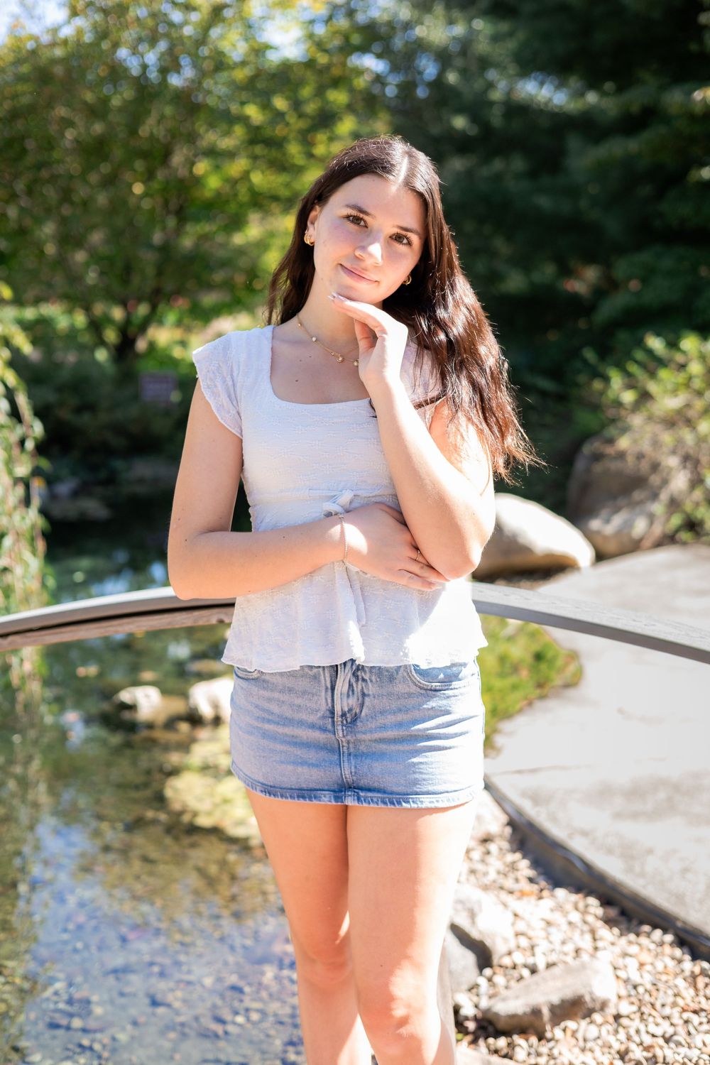 Young woman in white top and denim skirt poses thoughtfully by a serene garden pond on a sunny day.