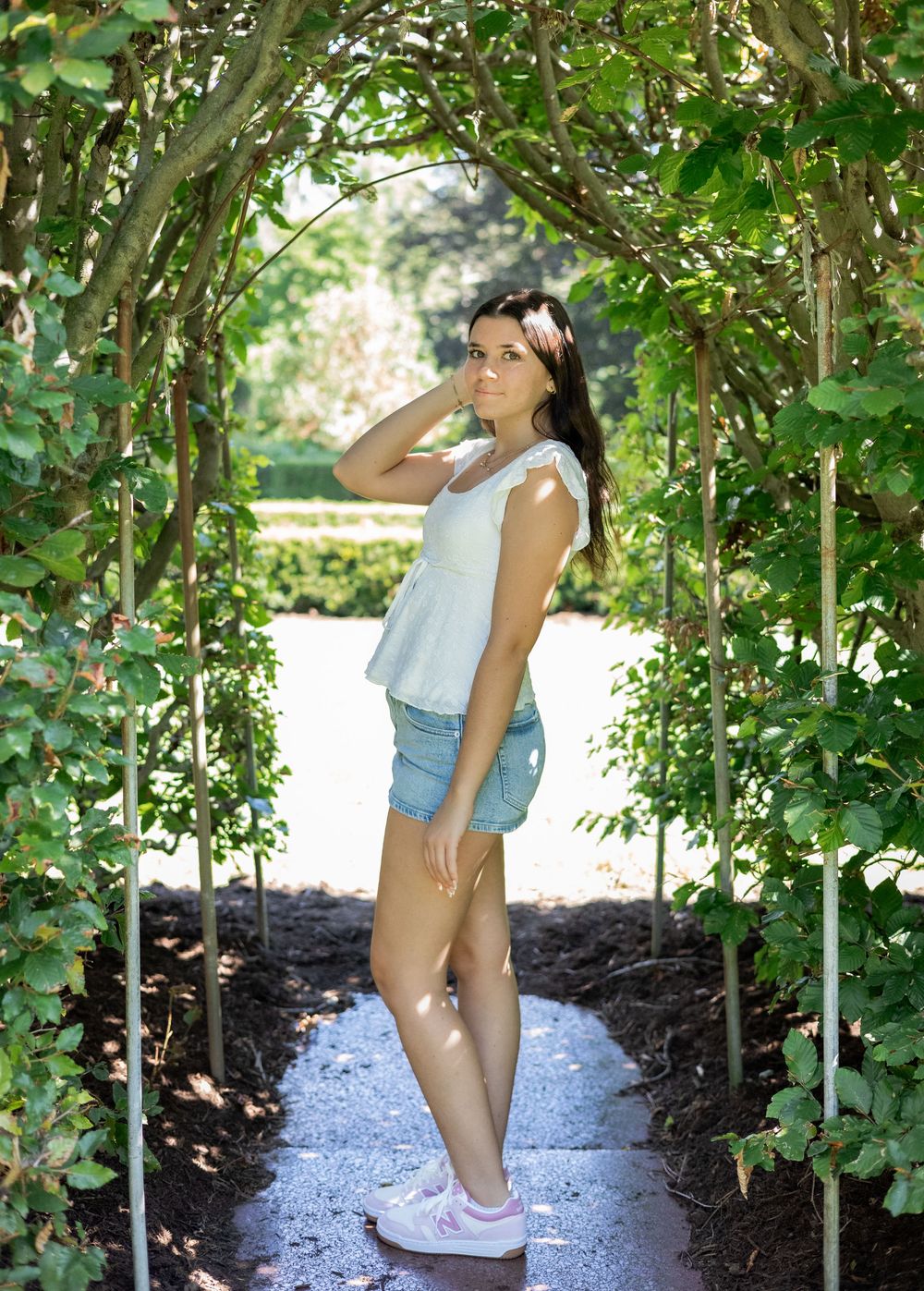 Young woman in white top and denim shorts poses under a lush green garden archway on a sunny day.